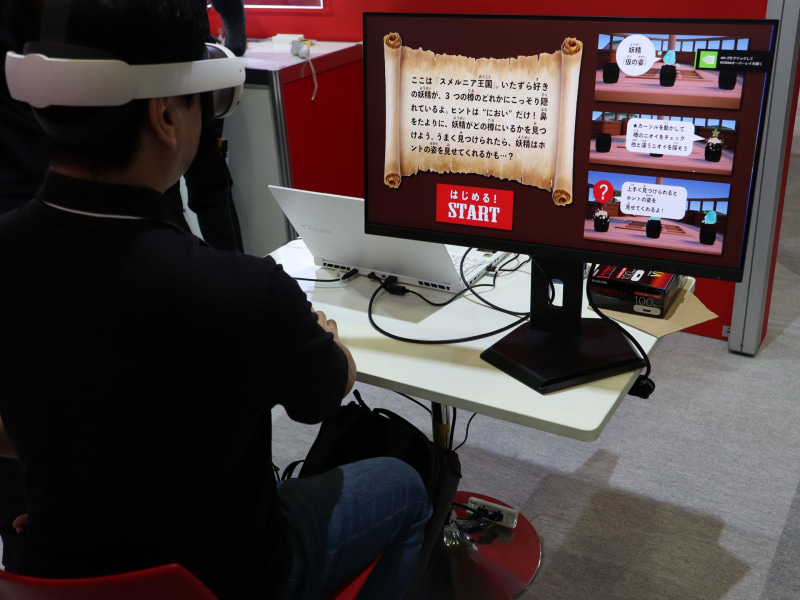 産業技術総合研究所のブース（<a href="https://www.ceatec.com/nj/exhibitor_detail_ja?id=36" class="strong bn" target="_blank">ホール4 ブース番号4H621</a>）には体験型の展示が多数。写真は、微量ガス制御による「小型ウェアラブル嗅覚ディスプレイ」とVR技術を連携させることで、人の嗅覚特性を取り込んだ高度な“匂い体験”の実現を目指す研究の展示。同ブースのそのほかの展示については、事前インタビュー記事<a href="https://internet.watch.impress.co.jp/docs/interview/2143553.html" class="strong bn" target="_blank">『「モアレ画像で“たわみ”測定」「座ったままで転倒リスク判定」「匂いが出るVR」――社会実装へ、産総研が本気の挑戦』</a>を参照してほしい