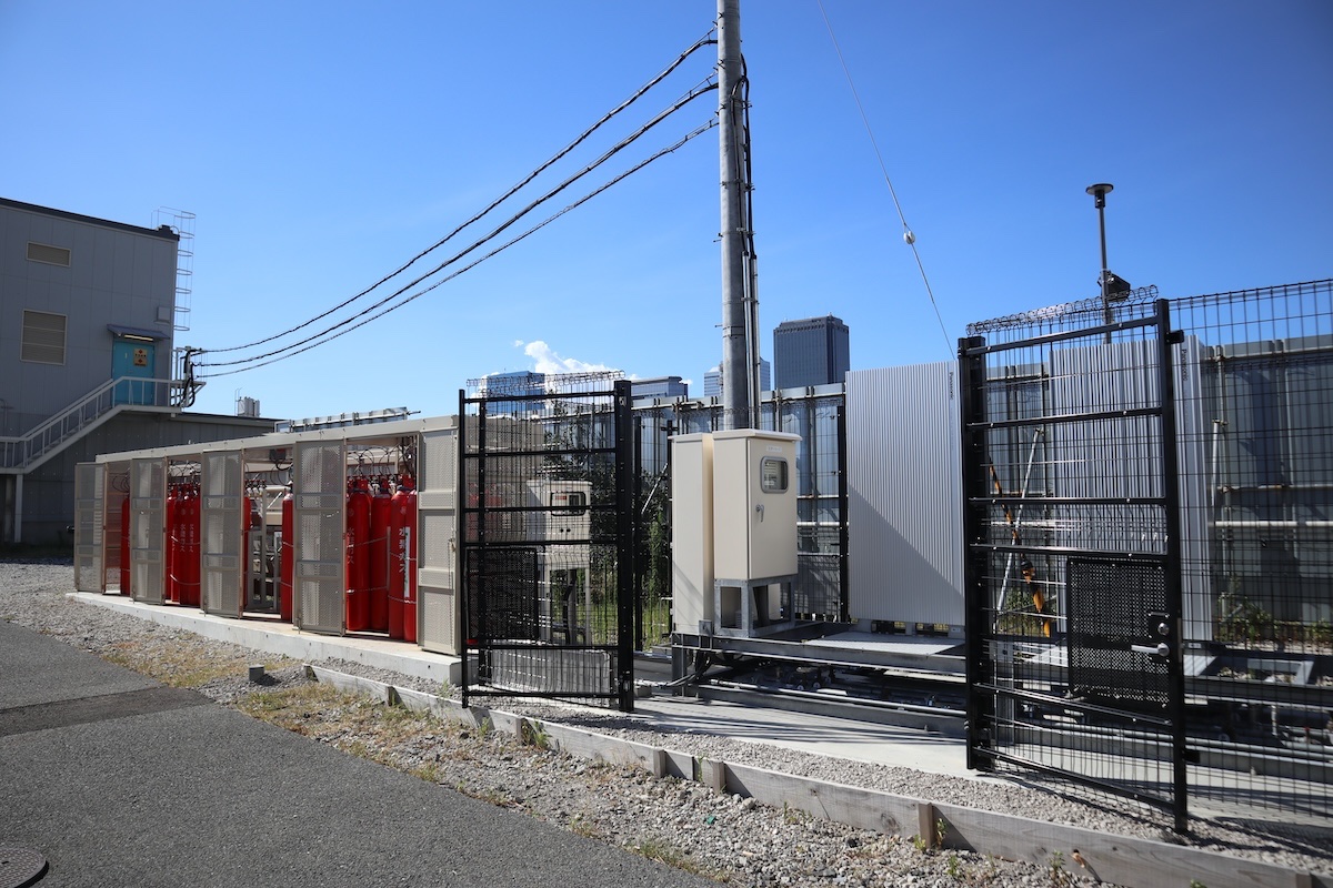 右側のタンスが燃料水素電池。太陽光パネルは壁の向こう側に設置されている。左側にある赤いボンベは水素貯蔵用タンク。その奥にある建屋に電線が張られ、建屋の中に昇圧用のキュービクル(変電設備)がある