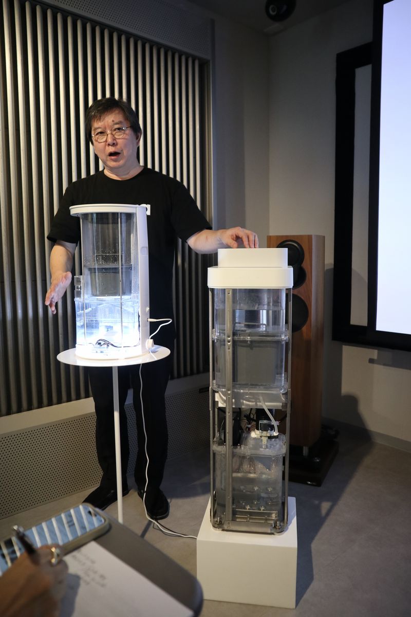 プルガティオの森久康彦 代表取締役。右側の大きな初代「moya Ultra」(プレハブ44畳まで)と小型化した「moya Craft」(プレハブ13畳まで)のスケルトンモデルでしくみを説明