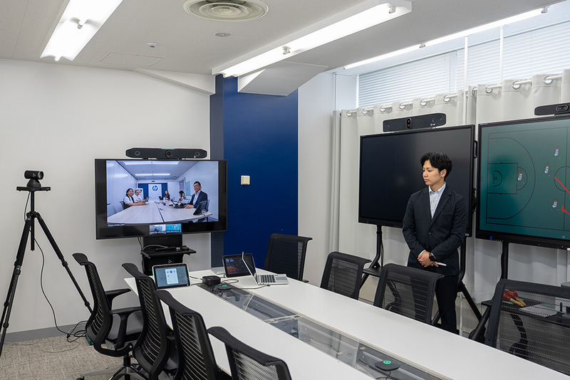 先生が遠隔授業をしているシーンを再現。手前の机の上にあるマイクと先生は少し離れているが、声だけがはっきりと届く。奥のディスプレイには授業を受ける教室を映すことも可能（実際は隣の会議室の様子を表示）。生徒の様子も先生は把握できる