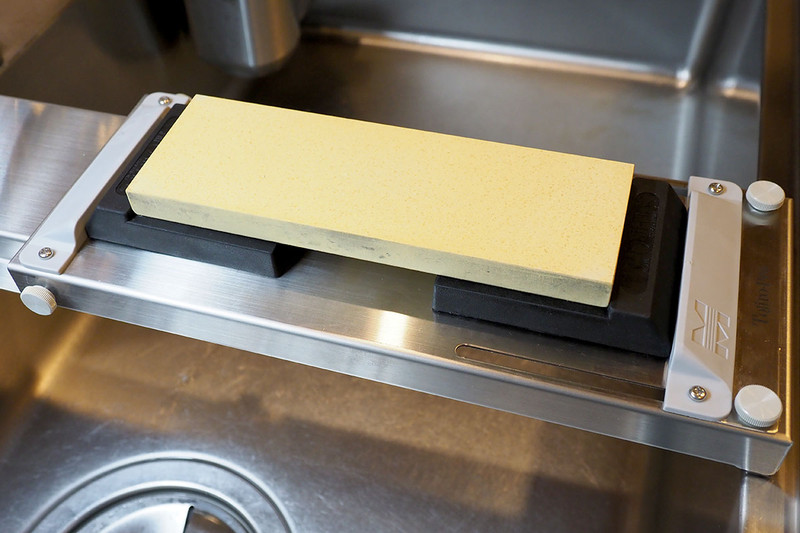 裏面にある、シンクのフチに引っ掛ける部分には滑り止め・傷付け防止のラバーがあります。机上など平面に置いて使うことも可能。砥石を固定する押さえ部分の高さは約13mmありますが、薄めの砥石だと固定できても「押さえ部分やその周辺のネジなどが手に当たりがち」だったりもします。その場合は写真のように市販の砥石台を置いた上で固定するという方法も。写真の黒い砥石台は、スエヒロの「砥石台 フリー SKG-42」（<a href="http://www.suehiro-toishi.com/toishidai/skg-42/" class="n" target="_blank">公式ページ</a>）です。