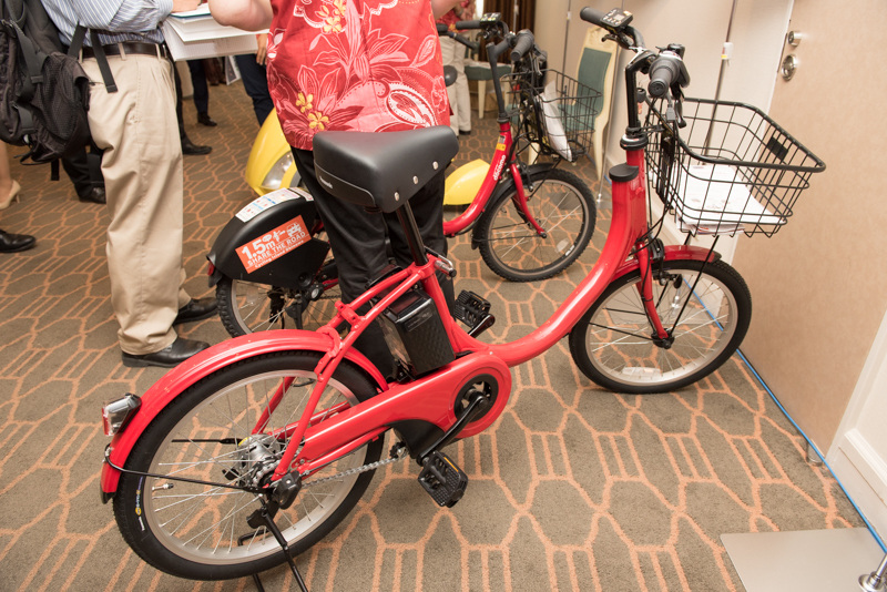 那覇市で導入が始まっているシェアサイクルは新型も展示。「ちよくる」など首都圏のサービスでも順次置き換わっていく予定という。写真はパナソニック製で、スマートフォンを充電できるUSBポートを備えているのが特徴。写真の車両は通信ユニットがまだ搭載されていない状態。ほかにブリヂストン製があり、こちらは回生ブレーキを装備するモデルになる。