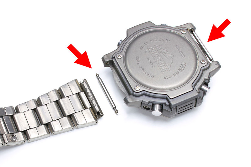 腕時計ホルダーに腕時計を装着するには、腕時計とバンドをつないでいるバネ棒（赤矢印の棒状部品）を外したり再度付けたりする必要があります。精密ドライバーでもバネ棒の付け外しは行えますが、滑りやすいので時計に傷を付けるリスクが高まります。バネ棒を外すための専用工具「バネ棒外し」（右写真・右下）があれば、より容易かつ比較的に安全にバネ棒を付け外しすることができます。なお、バネ棒なその名のとおり内部にバネが入っています。付け外しするとき、ビョーンと飛んで紛失したりすることがあるのでご注意を。