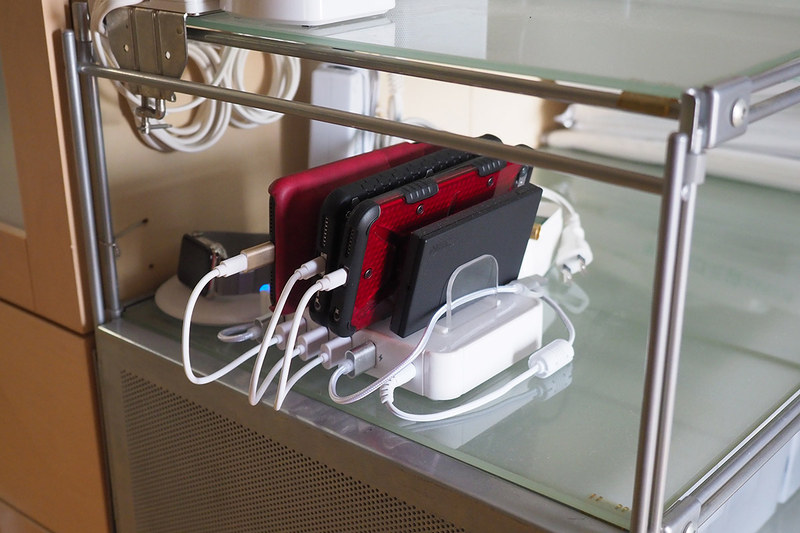 左写真は、単にUSB-ACアダプターと端末をつないで充電していた頃の様子（再現）。右写真は、ある充電スタンドを使って充電している様子。明らかに片付きますし、USBケーブルを短いものに交換すれば乱雑さ煩雑さも激減です！