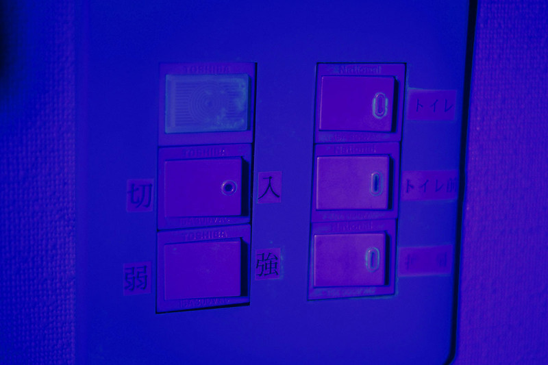 左は、可視光線とその付近の電磁波を説明した概念図で、虹色で示された範囲が「人間の目に見える波長の電磁波」。つまり目に見える光です。その外側の紫外線や赤外線は目に見えません。UVライトを当てた蛍光物質が光るのは、紫外線を受けた蛍光物質が（励起して）可視光線を発するからです。右の写真は壁のスイッチパネルにUVライトを当てた様子。UVライトに含まれる青紫の可視光線により、こんなふうに青っぽく見えます。少し薄緑っぽく見えるのは樹脂や皮脂に含まれる蛍光物質で、うっすらと発光しています。