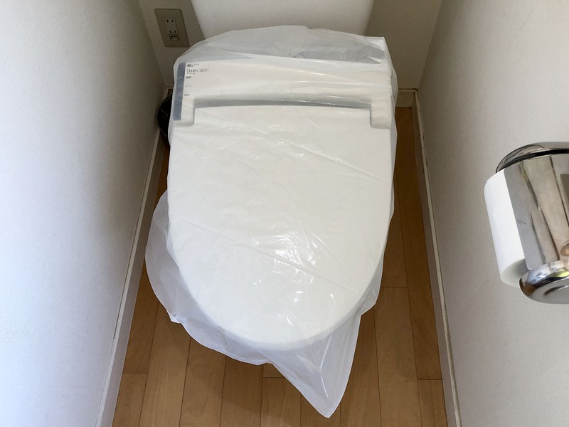 続いて、購入した温水洗浄便座に付属する新しいホースを取り付けます。じつは、同じブランドの製品だからか、古くから取り付けてあった配管類を流用できそうでした。しかし確認のため外してみると、古い配管類の接続部パッキンが少々痛んでいたり、錆がやや出ていたので、新しいものと交換することにしました。右写真は古い温水洗浄便座を取り外す前にビニール袋に入れている様子。取り外した温水洗浄便座からは僅かに水が漏れてくるそうなので、こうしました。取り外した温水洗浄便座はなるべく汚れを除去してから廃棄します。自治体の不燃ゴミ窓口へ持ち込んだら250円で処理してもらえました。