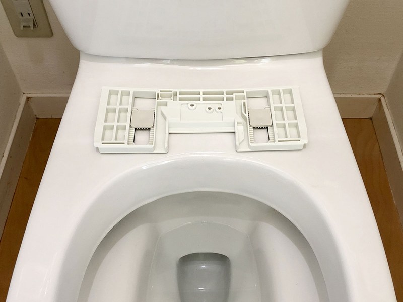 便座を外した様子。便器上部奥に見える2つの穴を使って便座を取り付けます。この便器の穴の間隔は140mm。多くの便器がこの穴間隔なのだそうです。これに合わせて温水洗浄便座も設計されているわけですね。右写真は、その穴に、新しい温水洗浄便座に付属する取付アダプター（本体脱着プレート）を装着した様子。便座をワンタッチで取り外せる便利なアダプターです。