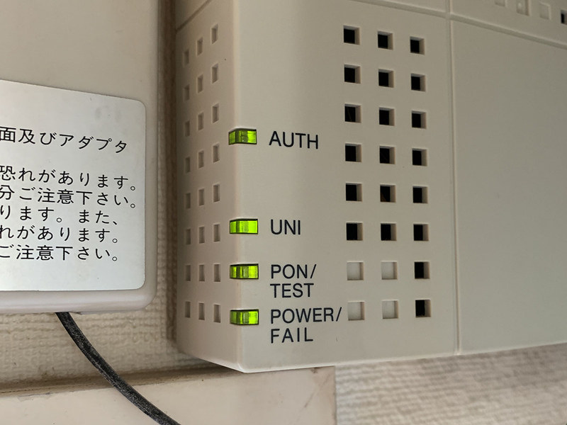 三菱電機製の「GE-PON〈M〉A GE-PON-ONU〈1〉〈2〉」というONUです。拙宅では窓の上の壁に取り付けてあります。ONU左下の4つのランプで動作状態がわかります。中央写真のように、4つのランプ全てが緑点灯（うちUNIランプは点灯もしくは点滅）の状態が正常。どれかのランプがオレンジや赤に点灯・点滅したり消灯したりし続けている場合、何らかのトラブルが起きています。右写真は拙宅の光回線ダウン状態のもの。AUTHランプが消灯したままですが、これは故障中か光回線運用準備中を示しています。