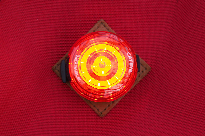 こちらはウェアラブルライトにもテールライトにもなる「SYNC WEARABLE」（<a href="https://www.cateye.com/jp/products/safety_lights/SL-NW100/" class="strong bn" target="_blank">公式ページ</a>）。曇天下での点灯イメージです。ライトのクリップを使い、バックパック上部のストラップ通しに装着。テールライトSYNC KINETICほどではありませんが、これも目立つ赤色ライトです。なお、SYNC KINETICのキネティックモード発光と連動させ、減速時に赤く明るく光らせることができます。