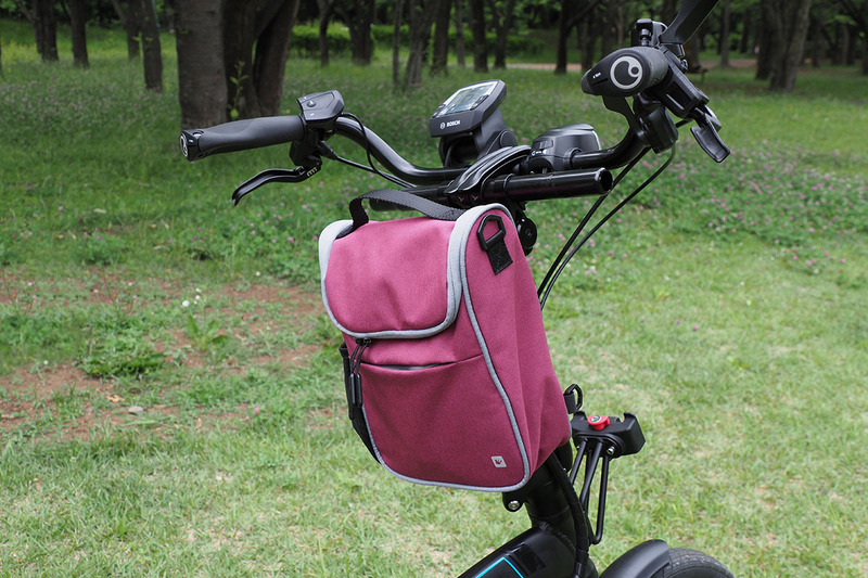 これはミニベロタイプのe-bike。装着しているアタッチメントは「キャディー（KR851）」という製品で、小径車のハンドルポストやシートポストへの取り付けに向きます。バッグがこちら向きになるように装着したら、モノの出し入れがラクで便利♪
