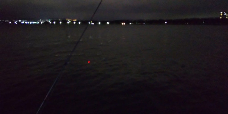 夜釣りの風景を撮影した3枚。周りに明かりがほとんどない、本当に暗い場所。手前に釣り竿、電気ウキが海面に浮かび、遠くに街の明かり。フラッシュオフ（左）、ライトオン（中）、フラッシュオン（右）。これはライトが一番良いと思うのですが