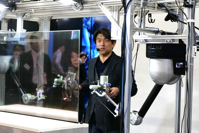 両手にコントローラーを握って操作する。手前の透明の板は半透過ディスプレイで、ロボットからのカメラ映像が表示されている