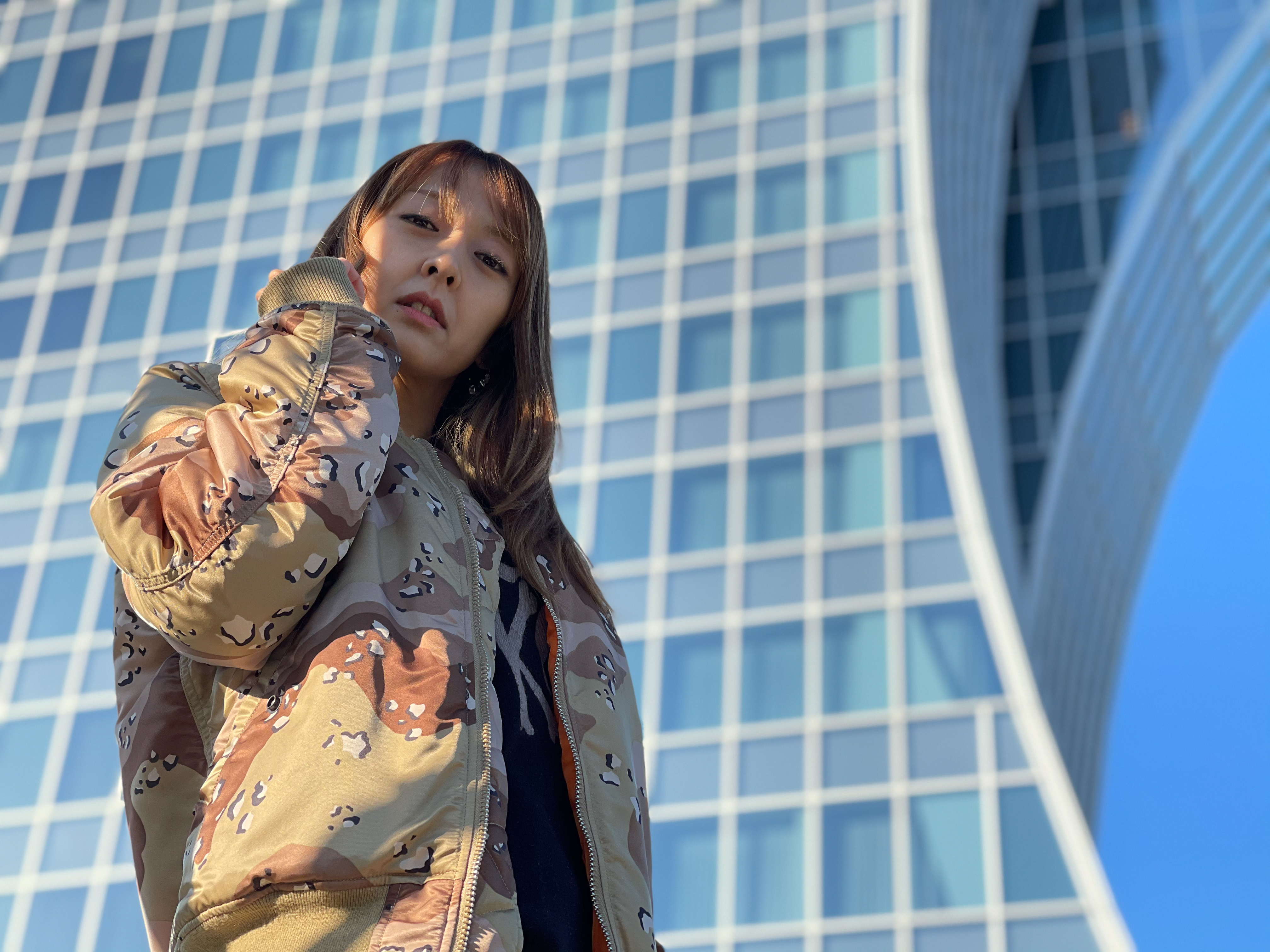 「iPhone 12 Pro Max」（左）、「Pixel 5」（右）。真新しいビルを背景にしてモデルをあおって撮ったカットだが、どちらも優秀な写りだ。やはりこのカットでも「iPhone 12 Pro Max」はモデルの顔を見た目より明るく写し出している。境界判定は肩口にかかる窓部分をキチンと判別できずボケていない