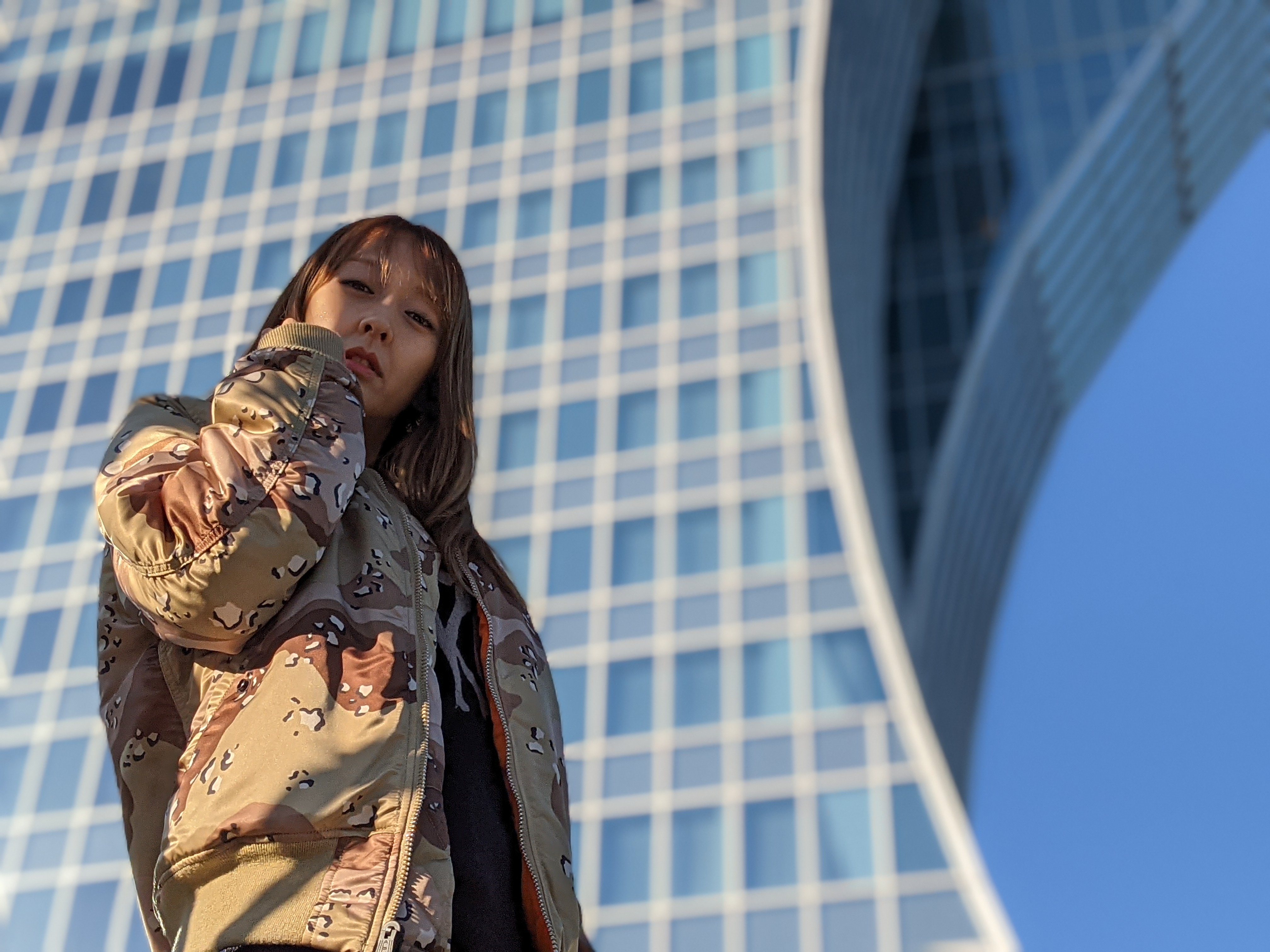 「iPhone 12 Pro Max」（左）、「Pixel 5」（右）。真新しいビルを背景にしてモデルをあおって撮ったカットだが、どちらも優秀な写りだ。やはりこのカットでも「iPhone 12 Pro Max」はモデルの顔を見た目より明るく写し出している。境界判定は肩口にかかる窓部分をキチンと判別できずボケていない