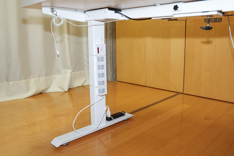 昇降デスクの左脚。こちらは別系統からの電源がつながっている。脚に固定した白いマルチタップから昇降デスク上の機器への給電を考えており、脚の下部に付けた黒いマルチタップからは昇降デスク上の（白いマルチタップ以外の）機器へは給電しない