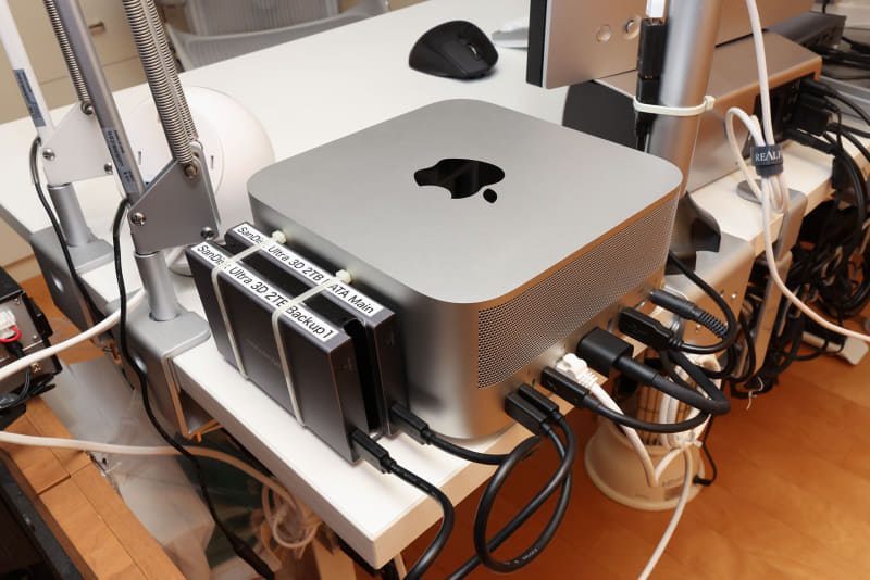 後ろから見た様子。Mac Studio導入を機会に、周辺機器接続インターフェースを整理。なるべくThunderbolt 4を使うようにしたら、少しスッキリした。左に見えるのはSSDだが、これも机に貼ってある。Thunderbolt 4ポートは、Thunderbolt 4（最大40Gb / s）/DisplayPort / USB 4（最大40Gb / s） / USB 3.1 Gen 2（最大10Gb / s）として使える