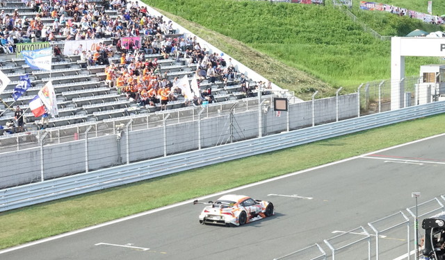 au TOM'S GR Supraとファンシートで応援する人々（オレンジ色の服装）。過去には全日本GT選手権時代に「au セルモ スープラ」というマシンもあり、KDDIとモータースポーツの歴史は古い