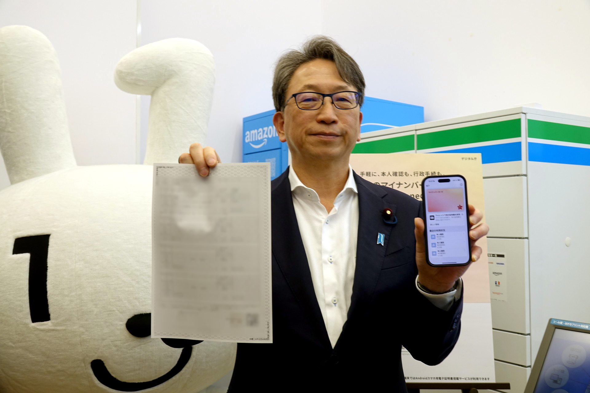 自身のiPhoneと住民票の写しを手にする平大臣