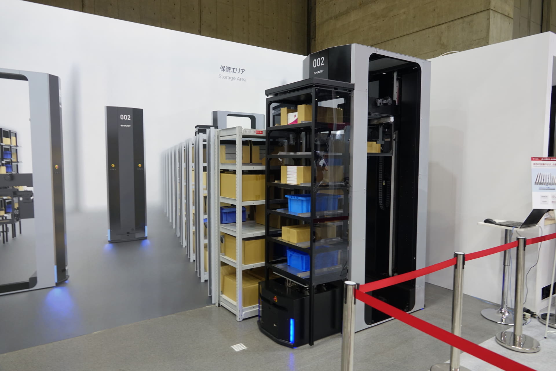 ピックアップするようす。奥のグレーの棚からロボット（手前左側）のアームが荷物を引っ張り出し、収納棚（手前右側の黒い棚）に移し替える