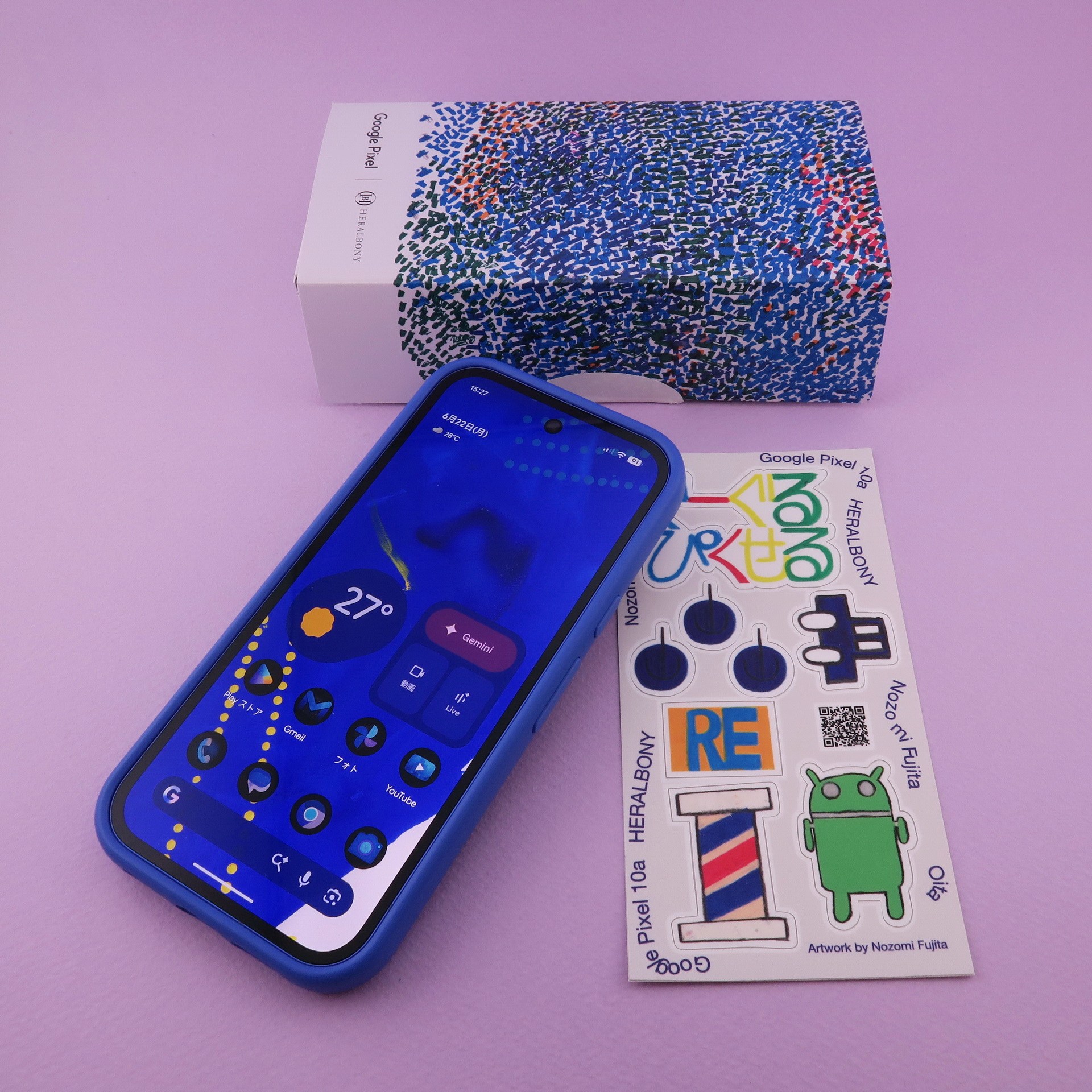 「Pixel 10a」の日本限定モデル「Isai Blue」は、標準カラーの内容に加え、本体の側面に装着するタイプのバンパー（写真内では端末に装着済み）、ステッカー（右）が同梱される。パッケージ（後ろ）もオリジナルデザイン