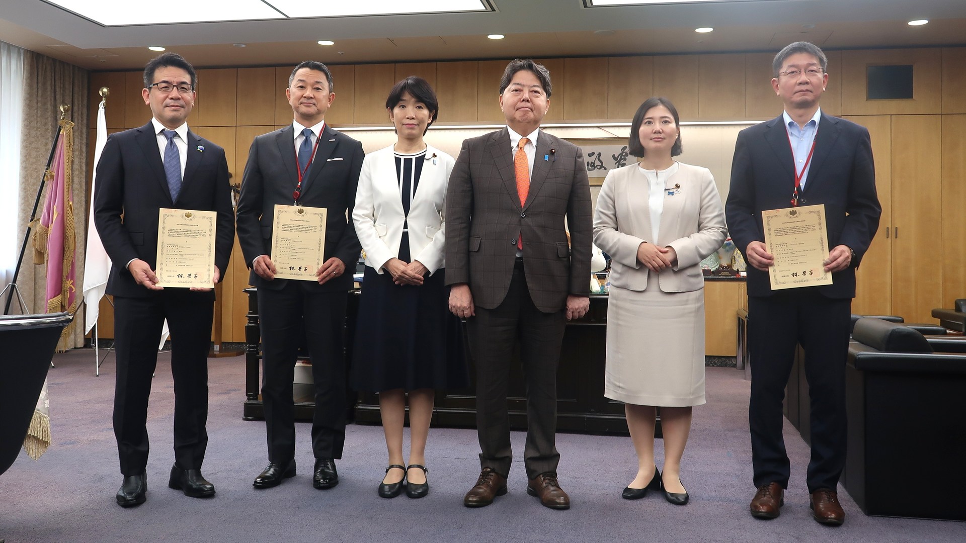 7月15日に総務省で行われた認定式の模様。左から、JTOWER代表取締役社長CEOの田中敦史氏、NTTドコモ代表取締役社長の前田義晃氏、堀内詔子総務副大臣、林芳正総務大臣、向山淳総務大臣政務官、ハイテクインター代表取締役社長の旦尾紀人氏。なお、全国枠（25.8～26.2GHz）はNTTドコモが、地域枠（26.8～27.0GHz）はJTOWERとハイテクインターが落札していた