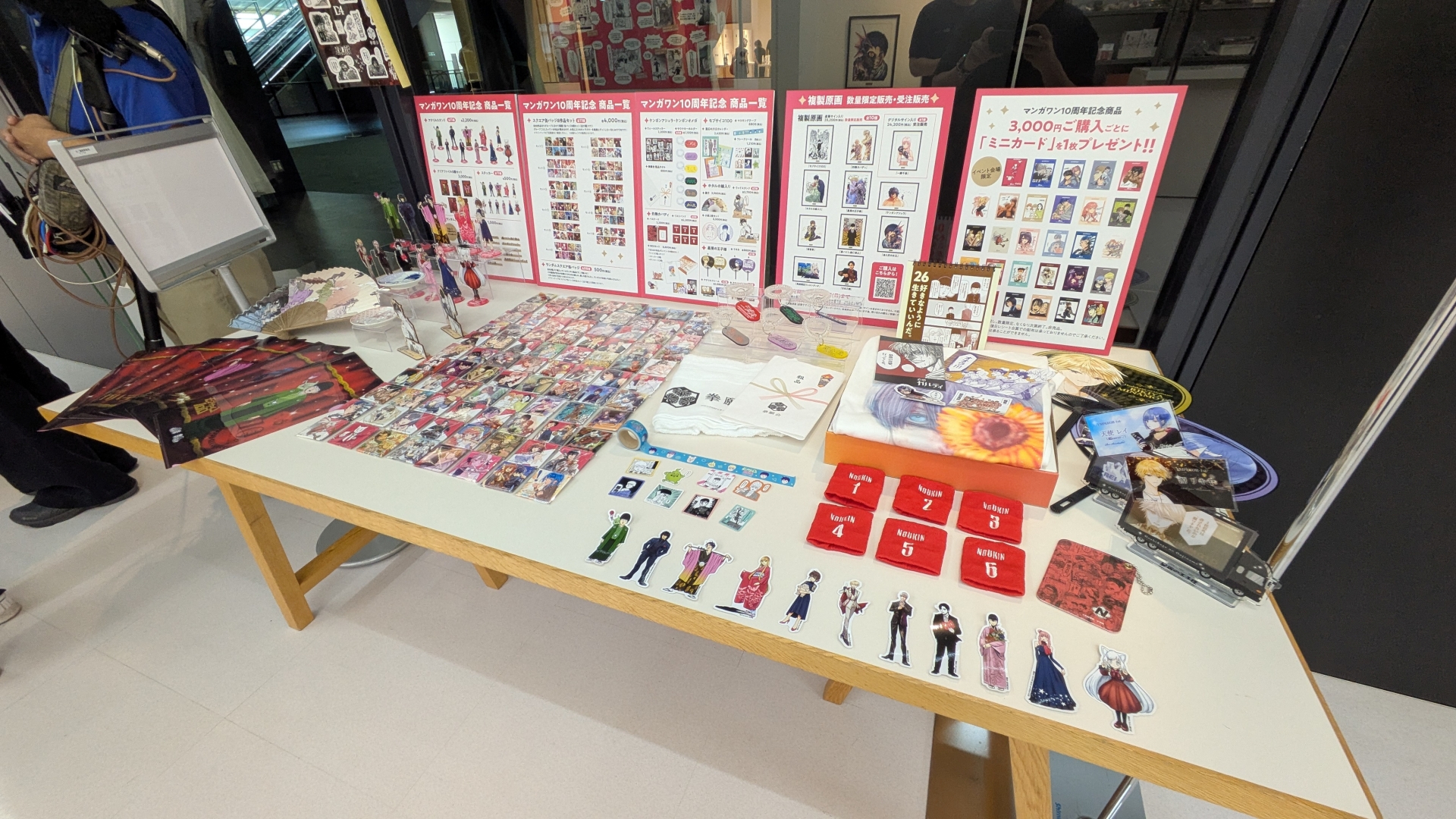 物販コーナーのお品書きと展示。缶バッジやアクスタといったアイテムのほか、数量限定で複製原画の販売（デジタルサイン入りの「一勝千金」のみ受注販売）なども行なわれる。複製原画は展示も行なわれていたが、思わず「これが買えてしまうのか……」と思ってしまうほどの出来栄えだった