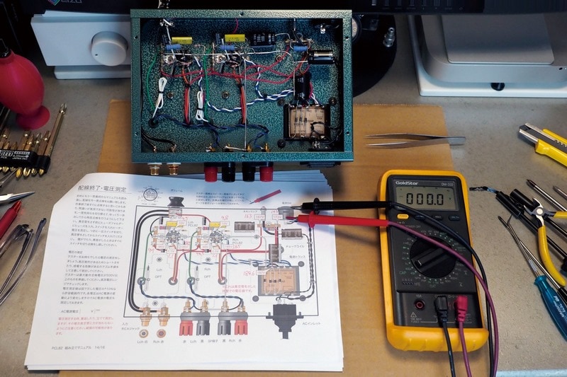 ここでテスターの出番である。真空管を挿し込んで裏返して電源ケーブルをつなぐ。テスターの黒棒をスピーカー端子のマイナス側に固定。赤棒は先端以外はテープで巻いて絶縁。電源を入れて電圧を測定する。この際、ゴム手袋を着用のこと