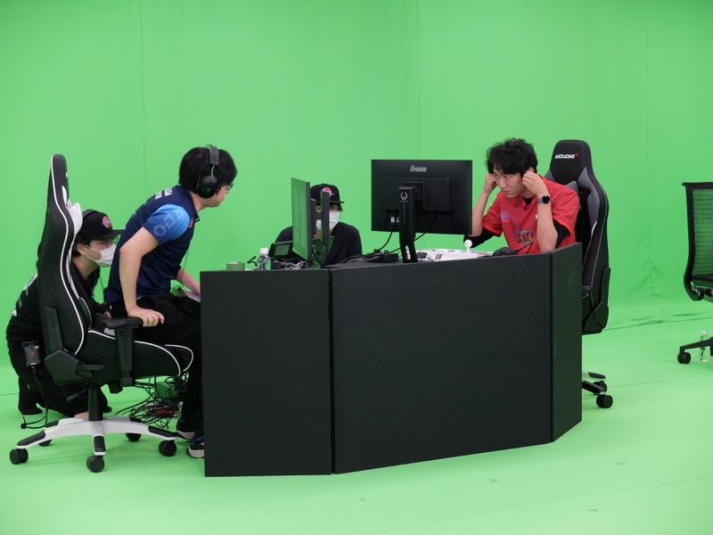 実際の会場の様子。チームごとに同じPCを使い、コントローラーとイヤフォンのみ各選手の物を接続してオフライン対戦を行なう。背景は合成のために緑一色の背景だ