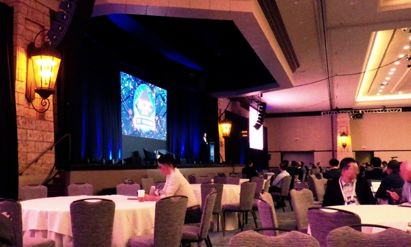 キーノート講演の会場風景。円形のテーブルに着席する。大型スクリーンが前方と後方の壁面に3枚ずつ配置してあり、大部分の座席からスクリーンを眺められる。現地時間2025年5月28日午前8時過ぎに撮影