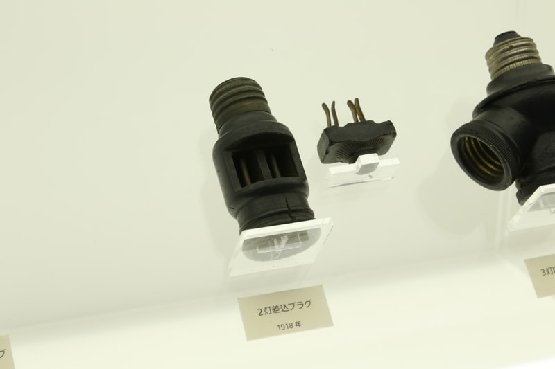 1918年(大正7年)「二灯用差し込みプラグ」。電球ソケットの横に小さい歯が付いた部品を差し込むと、ここから電気が取れるアダプタ。形状は現在のコンセントに近い。これはちょっと工作が必要なのでマニア向けで、後に「二灯用クラスター」が発売された