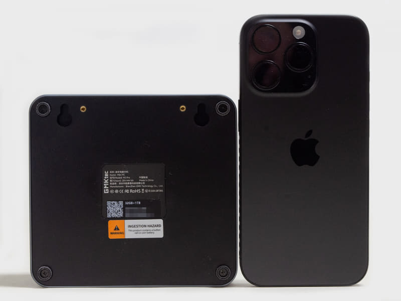 裏面とiPhone 16 Proの比較。四隅にゴム足と写真下側にVESAマウンタ用の凹み。一般的なミニPCと比較して少し小さいだろうか