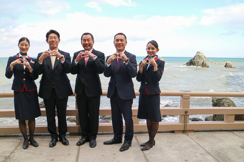 夫婦岩の前で、手でハートの形を作って記念撮影。写真左から、三重県四日市市出身のCA（客室乗務員）の真弓沙穂氏、鈴木健一市長、大西賢会長、鈴木英敬知事、三重県四日市市出身のCAの山本陽子氏