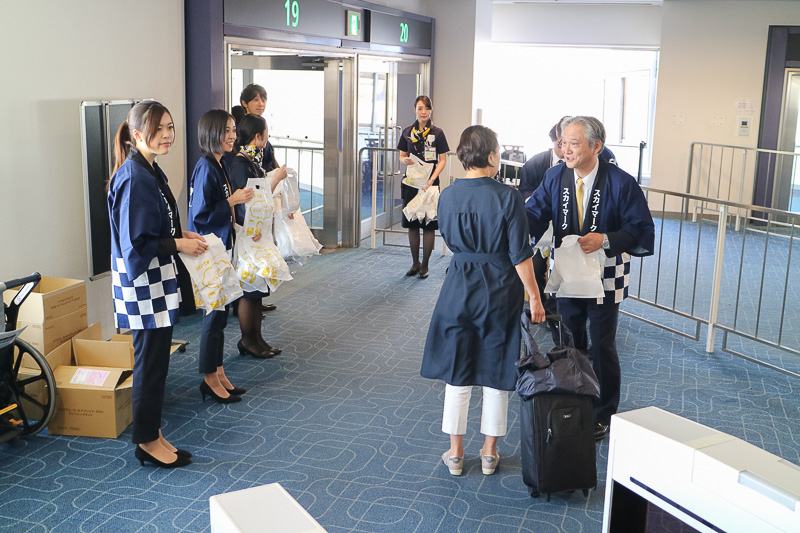 羽田空港 第1ターミナル19番ゲートにおいて、新千歳（札幌）行きSKY711便の搭乗客にスカイマーク株式会社 代表取締役社長の市江正彦氏や、同社スタッフが搭乗客にプレゼントを手渡した