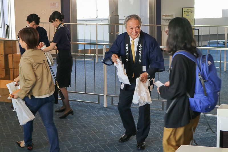 羽田空港 第1ターミナル19番ゲートにおいて、新千歳（札幌）行きSKY711便の搭乗客にスカイマーク株式会社 代表取締役社長の市江正彦氏や、同社スタッフが搭乗客にプレゼントを手渡した