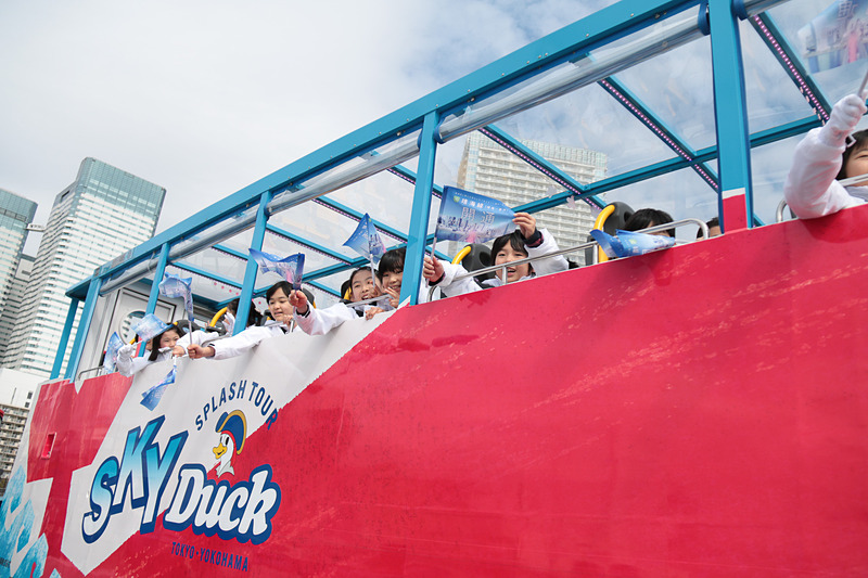 地元小学生や関係者らを乗せた2台のバスが通過