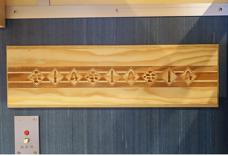 1号車（写真左）、2号車（写真右）の窓側壁面に貼られた装飾。1号車は隠岐の黒松（島根県隠岐の島町）、2号車は智頭杉（鳥取県智頭町）を用い、絵柄も出雲、因幡の神話にちなんだものとしている