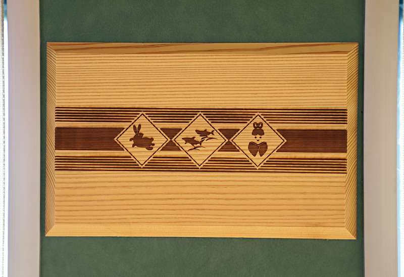 1号車（写真左）、2号車（写真右）の窓側壁面に貼られた装飾。1号車は隠岐の黒松（島根県隠岐の島町）、2号車は智頭杉（鳥取県智頭町）を用い、絵柄も出雲、因幡の神話にちなんだものとしている