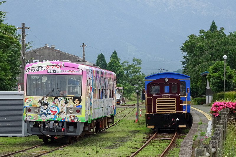 南阿蘇鉄道のトロッコ列車の旅へ。写真は高森駅に停車する「がんばれクマモト！ マンガよせがきトレイン」（左）と、トロッコ列車「ゆうすげ号」（右）