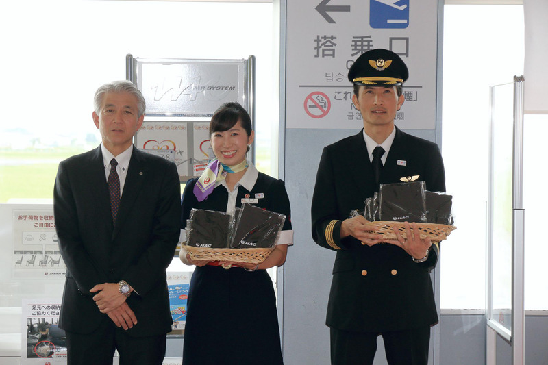 桑野氏とパイロットの岡田耕造氏、CAの酒井葉美氏によって記念品「鶴丸ロゴ入りHACオリジナルトートバック」が搭乗客に贈呈された