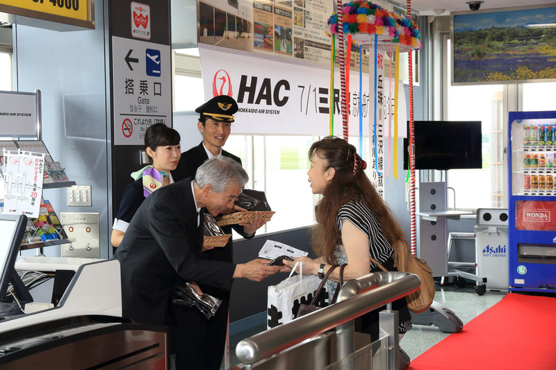 桑野氏とパイロットの岡田耕造氏、CAの酒井葉美氏によって記念品「鶴丸ロゴ入りHACオリジナルトートバック」が搭乗客に贈呈された