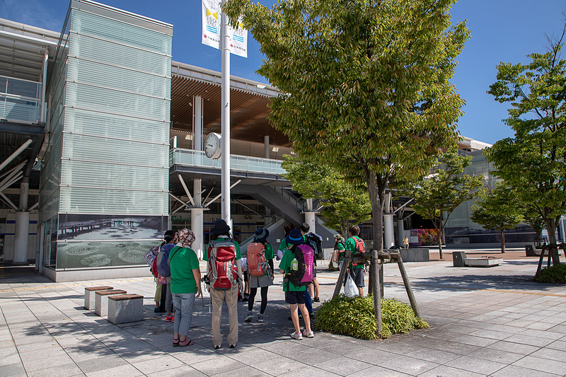 新潟駅前で参加者を待つ荻田氏とスタッフ。すでに汗ばむ気温なので木陰をキープ