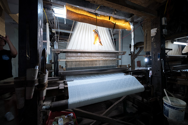 こちらは機械織りで、発動機の音と、横糸が行き来する音が工房に響き渡っていた。機械の上に垂れ下がった木の板によって模様を作るそうだ