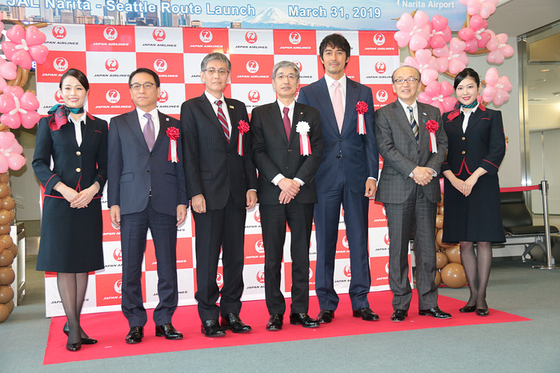 JALのCA（客室乗務員）が挟んで、左から成田国際空港株式会社 取締役営業部門長 荒川武氏、国土交通省 東京航空局 成田国際空港長 石井靖男氏、日本航空株式会社 代表取締役社長 赤坂祐二氏、JAL国際線イメージキャラクターを務める俳優の阿部寛さん、成田市長 小泉一成氏