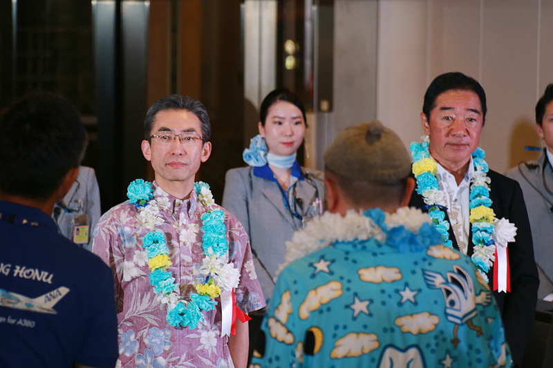 搭乗者をゲートで迎え一礼する平子社長と石田成田空港支店長。ディスプレイにはラニの画像も表示。フライト時間は7時間20分、現地は曇り、気温は23℃（摂氏）と表示されていた