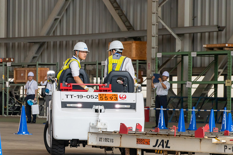 トーイング・トラクターでコンテナ・ドーリーをけん引しながらバック走行。運転者は進行方向、補助者は死角になる場所を確認しながら進む