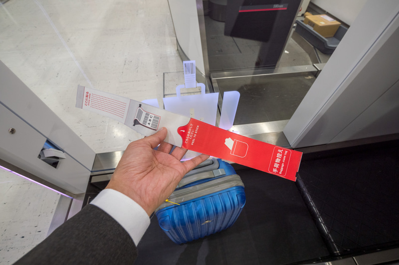 手荷物タグの貼り付け。剥離紙を剥がして取っ手などに巻き付け、シールを貼る。剥離紙側に手荷物控えが残るようになっている