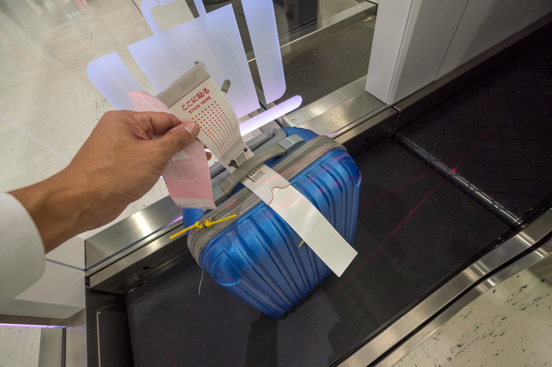 手荷物タグの貼り付け。剥離紙を剥がして取っ手などに巻き付け、シールを貼る。剥離紙側に手荷物控えが残るようになっている