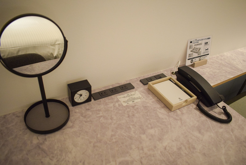 デスク回り。電話は内線のみ。メモ用紙＆ボールペン、アラーム付き時計、鏡とシンプル。鏡の土台はトレー状になっているので、アクセサリー類を置くのに便利。コンセントは3口、LANのコネクタもここにある