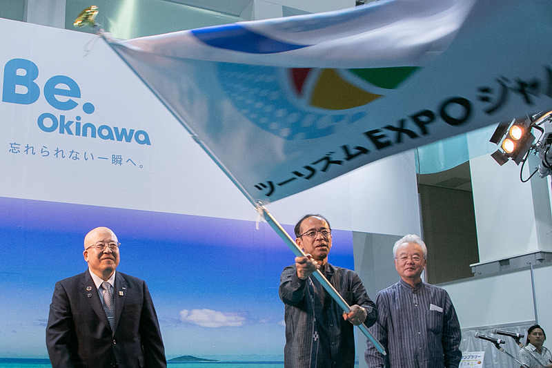 2019の会場で行なわれた「ツーリズムEXPOジャパン 2019→2020 ハンドオーバーセレモニー」。旗を振るのはOCVB（一般財団法人沖縄観光コンベンションビューロー）会長の下地芳郎氏