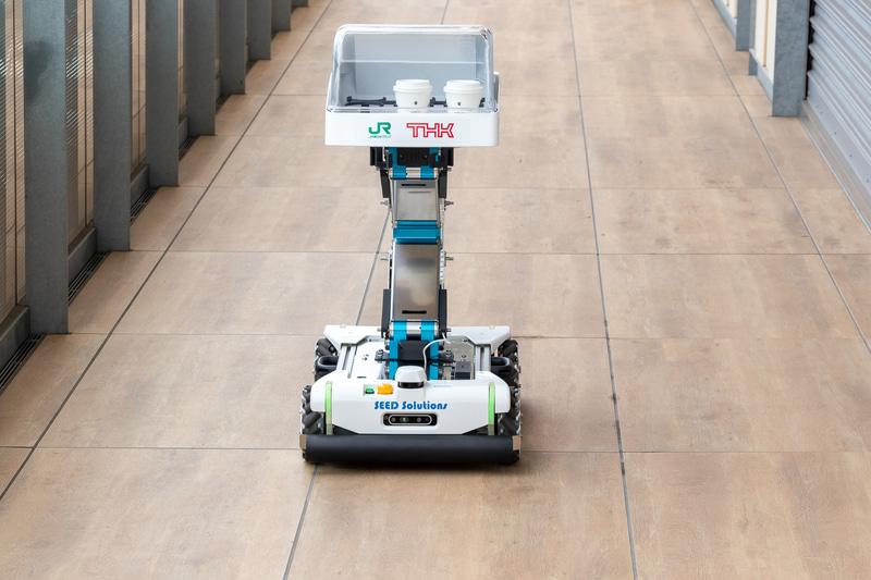 Lifter付きSEED-Mover。開発元はTHK。軽食や飲料の運搬を想定したパーソナルサイズの小型配膳ロボット。この日は2階の通路から3階のミーティングスペースの前までホットコーヒーを運搬。フロア移動に使うエレベータは自動で乗り降りする（エレベータの操作は手動）